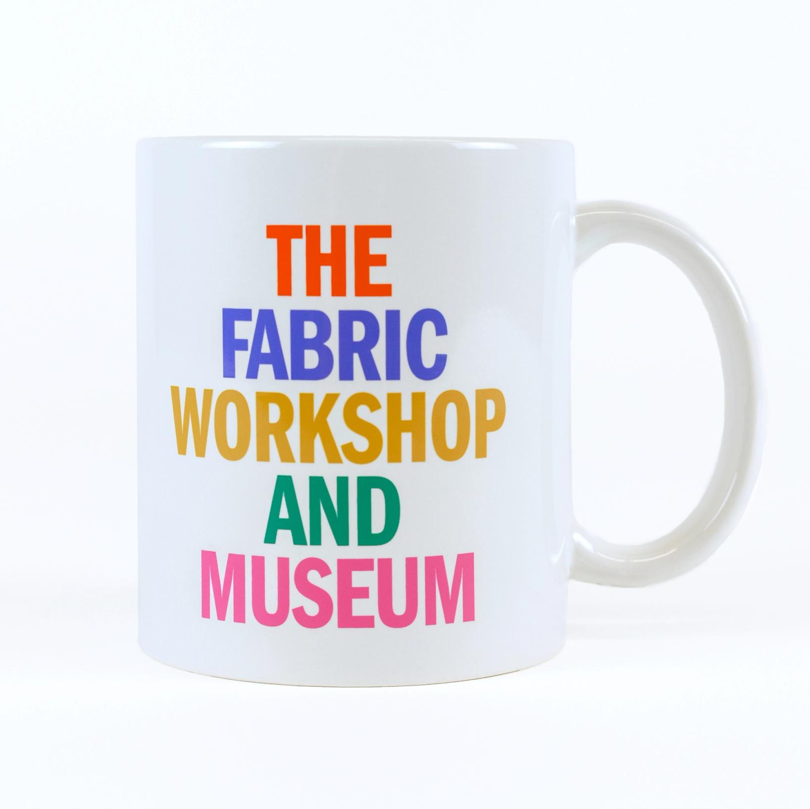 A cream colored mug against a white background says "THE FABRIC WORKSHOP AND MUSEUM". Each word is stacked and in a different color, red, blue, golden yellow, green, and pink.
