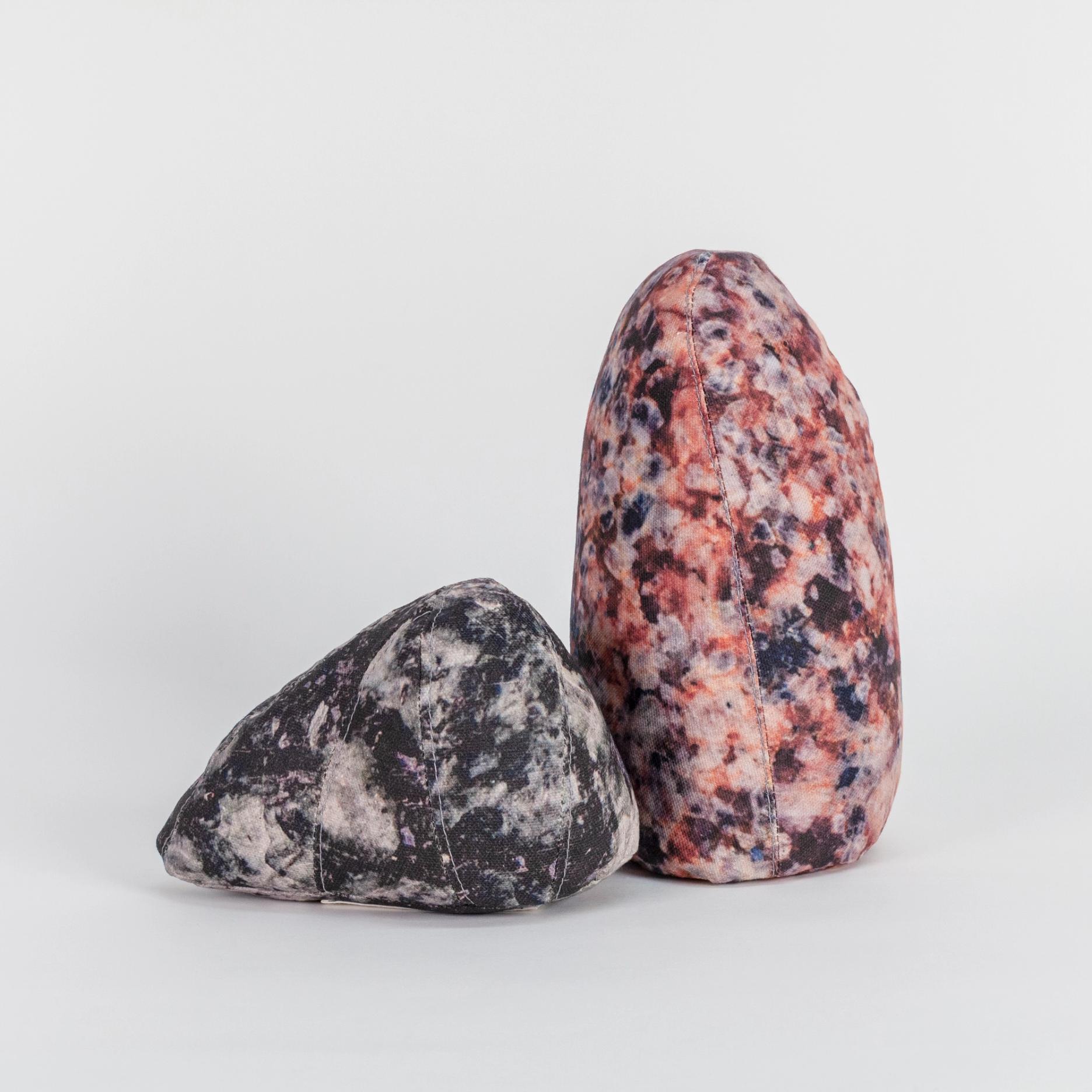 A photograph of two small soft sculptures covered with patterns that resemble polished stone of different hues. One sculpture is short and triangular where the other one is tall like an obelisk with a rounded top.