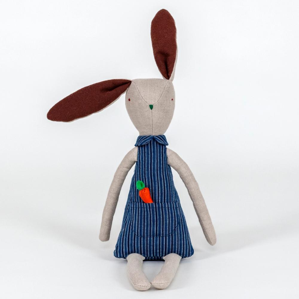 A photograph of a plush rabbit made of light beige woven fabric characterized to have a pointy nose, triangular shaped body, and long thin arms and legs. The body of the rabbit is made of a primarily blue patterned fabric and includes a pocket with a removable plush orange carrot.