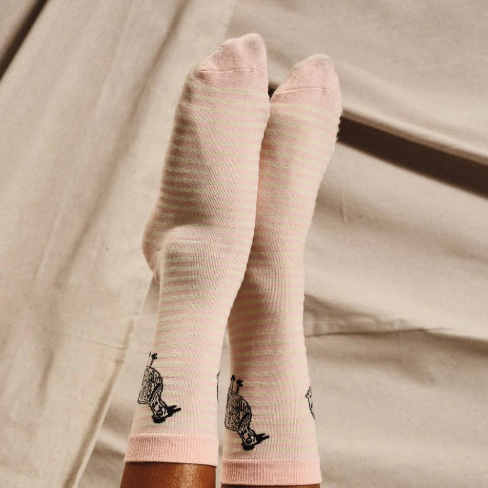 A person modeling a pair of light pink and white striped knit dress socks with a black illustrated Kuku bird on the left and right sides of the ankle of the socks.