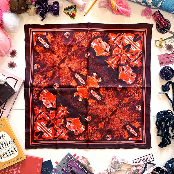 A square scarf with an abstract red pattern that is mirrored along the diagonal corners and bordered with thick maroon and thin pink stripes is placed at the center of a photograph. Surrounding it on all sides are an array of objects and materials from the artist Aimee Koran, including sculptures, patches, books, and fabric remnants.
