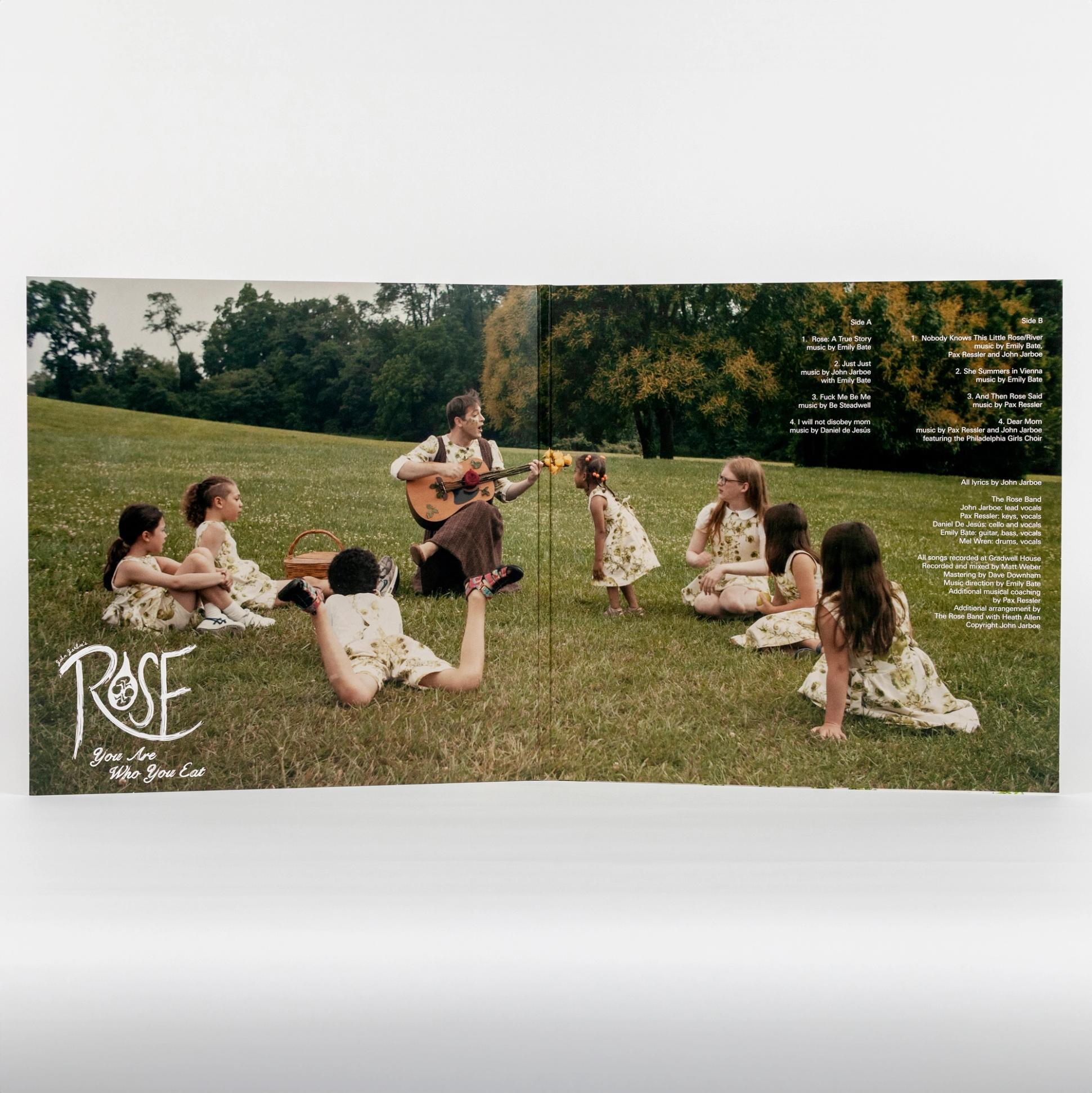 A photo of a record cover open to the inside spread. The spread features a single photograph spanning both sides. The image is of John, the artist, playing guitar and singing to a group of seven children on a grassy hill. The bottom left of the left side features "The Rose Garden" title graphic. The top right of the right side features the playlist for sides A and B of the vinyl record.