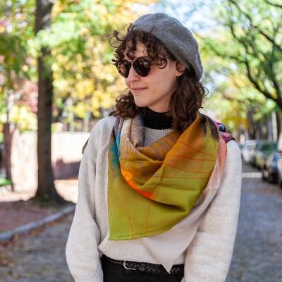 A person is wearing the scarf draped over an oatmeal sweater. They are wearing sunglasses and a grey beret, and you can see a cobble stone city street lined with trees and fall foliage behind them.