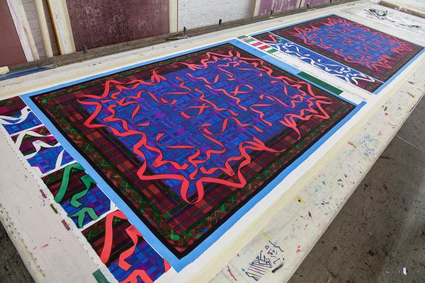 Two patterned textile designs recede in perspective from the bottom left of a photograph to its top-right. Dark red and green borders surround a deep blue at the center of the designs. Overtop is are mirrored ribbons of magenta.