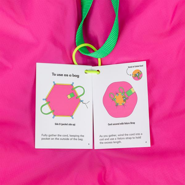 Instruction booklet against a bright pink fabric background showing diagrams for using a hexagonal picnic blanket, with steps for laying it flat and cinching the outlining cord to convert the blanket into a carrying bag.