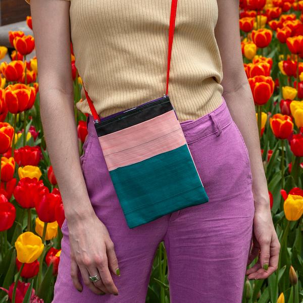 A model wears a small flat bag on her hip. The bag features stripes of teal, light pink and black, and has a red strap. The person is standing in a park with red and yellow tulips behind her.