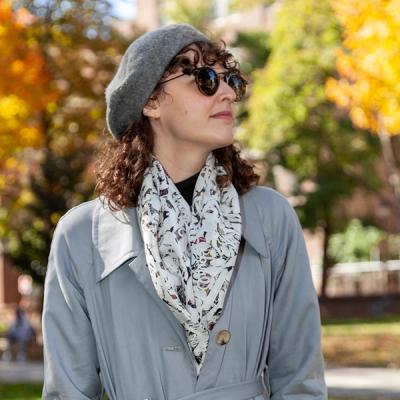 A person with curly brown hair, sunglasses, and a grey beret is wearing the scarf loosely tucked into a grey trench coat. In the background, out of focus, you can see a city park in autumn.