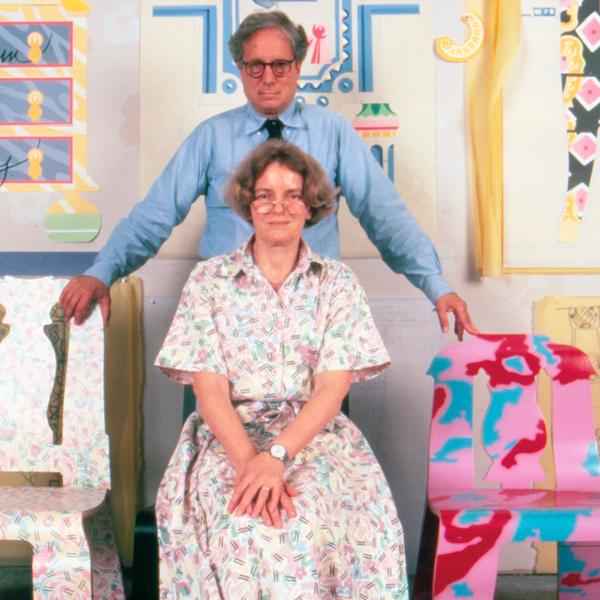 A portrait of a man and woman posing for the camera. The woman is wearing a faintly colored patterned dress with a color and sleeves to her elbows. She is seated with hands folded on her lap. The man, who is standing behind her with arms outstretched to rest his hands on a set of chairs on each side of them, is wearing a long sleeve blue dress shirt and a black tie. Behind them is a wall of painted pop designs that suggest furniture.