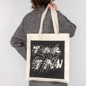 A person wearing a grey coat is facing away from the camera against a white backdrop. Slung over their shoulder in their right hand and facing the viewer, is a cream colored canvas tote with a black ink square screenprinted over the front of the bag. The negative space in the print has crosshatched lines intersecting with thick handwritten text that reads "TAR" on the top line and "TAN" below.