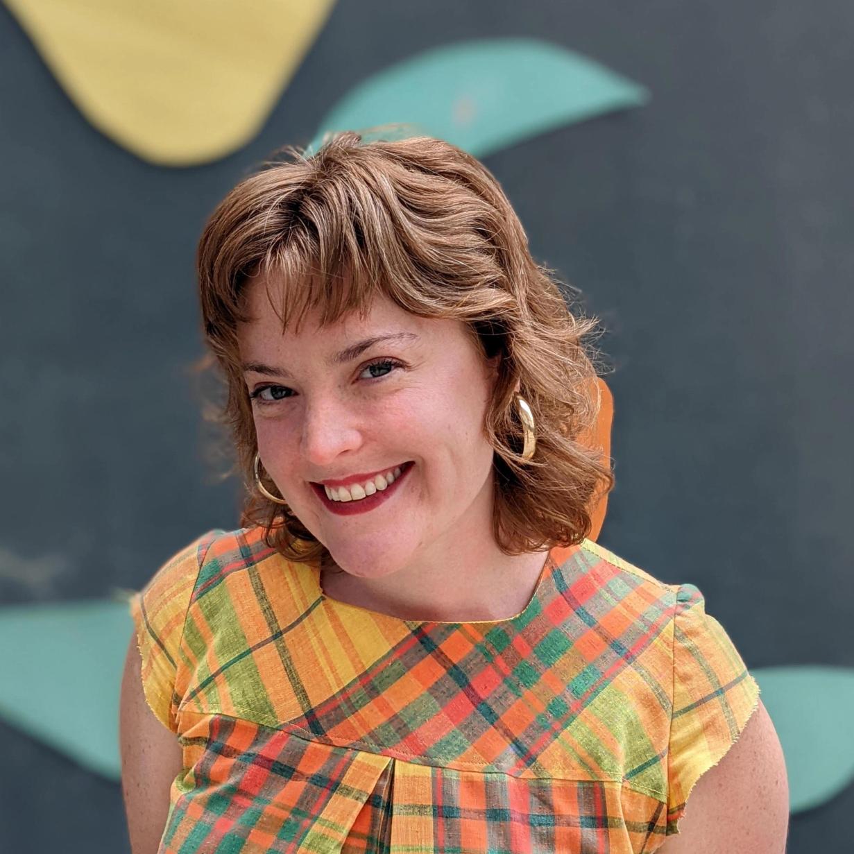 The artist, Clara Harlow wearing a plaid shirt, smiling and posing in front of a gray background.