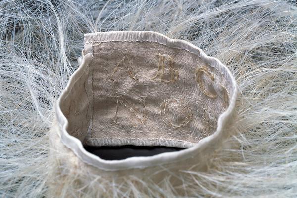 A photographic detail of a linen collar surrounded by an atmosphere of blond horse hair. The interior of the collar shows two rows of stitched letters: on the top is A, B, and C; on the bottom is N, O, and P.