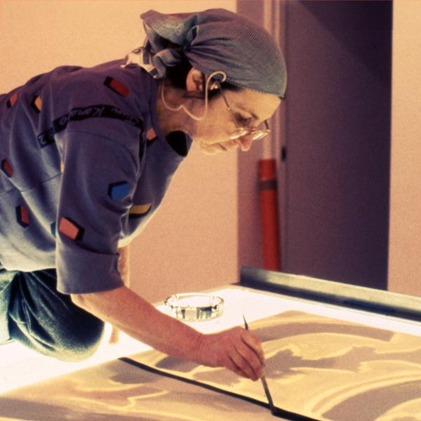 A photograph of a white woman artist leaning over as she paints on a surface illuminated by a light table. Wearing a headscarf and glasses, she is cast in yellow light.