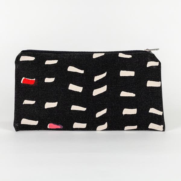 A canvas pouch, longer than it is tall, sits upright with a zipper resting on the top-right. Its design features a pattern of off-white dashes over a black ground. Two of the dashes are partially filled in with red.
