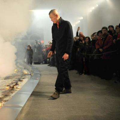 A male artist watching his performance piece comprised of a stainless steel trough of silk charmeuse fabric with gunpowder scattered on top that has been lit on fire and is smoking. The performance is taking place indoors in a gallery and there is a large roped off crowd behind the artist.