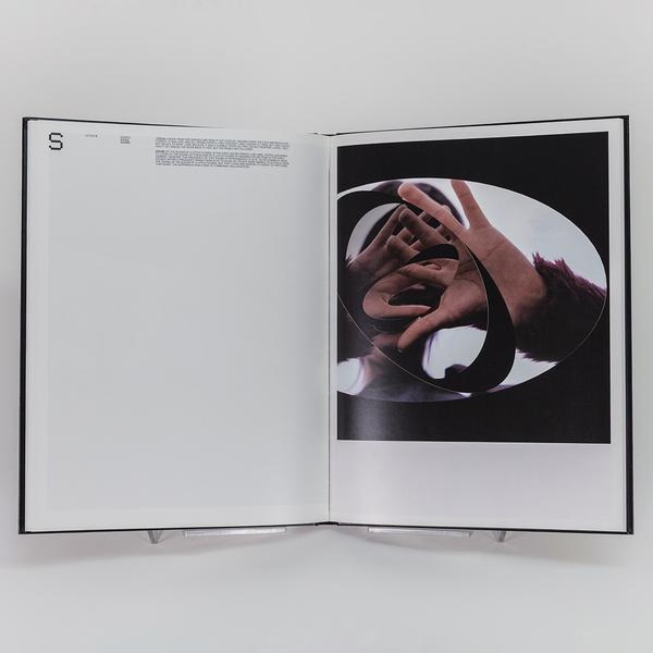 A photograph of an open book. The left page is marked with an "S" in the top left corner. the right page shows a photograph of a person making an oval shape with both thumbs and pointer fingers. The oval shape is echoed as the shape of the frame of the photograph.