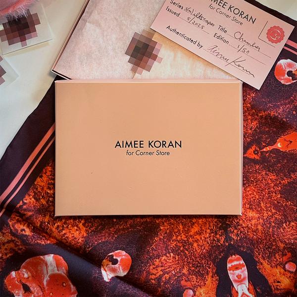A rectangular beige box lid that reads "Aimee Koran for Corner Store" is centered over a red scarf with a mottled texture. Above it is tissue paper  adhered with a pixelated nipple sticker and a pink certificate of authenticity.