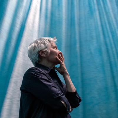 A photograph of a woman with short gray hair with her left hand resting on her mouth and chin as she gazes upward. She is surrounded by heavy fabric cast in blue light.
