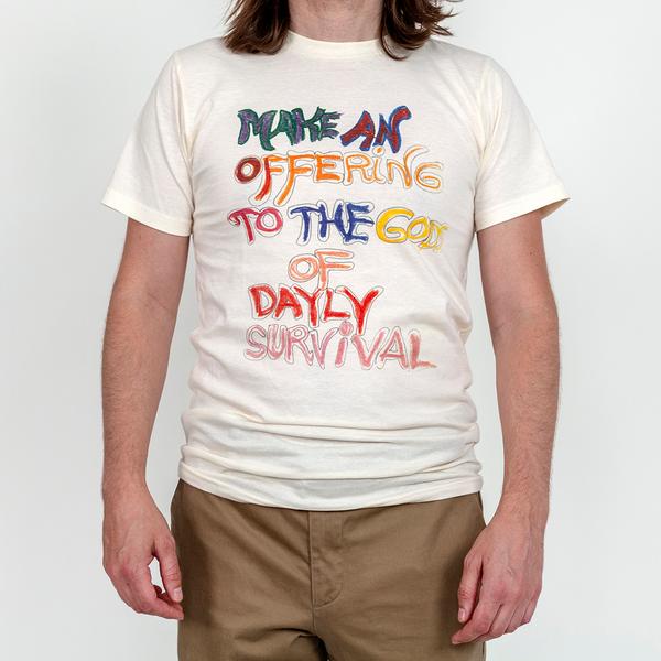 A model wears an off-white t-shirt that reads in a hand-drawn message, "Make An Offering to the Gods of Daily Survival." The word daily is spelled d-a-y-l-y. Each word is colored in with varying colors, including some individualized letters.
