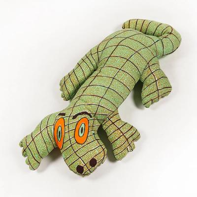 A photograph of a large plush toy resembling an alligator, printed in light green with dark brown lines in a grid across its body and small, rectangular orange scales. It has four thick legs with rounded toes and a curved tail. Its concentric ovular eyes include outer rings of orange, then light teal, and dark brown pupils. Its dark brown eyebrows and nostrils further punctuate its expression.