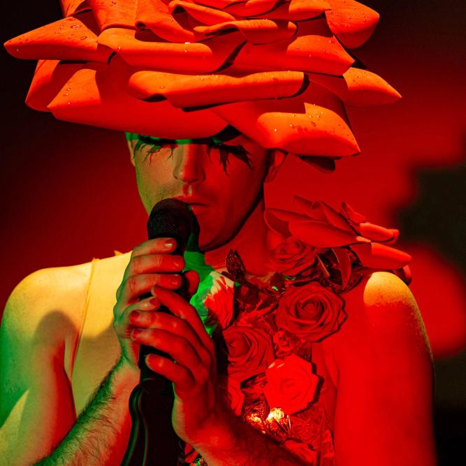 The artist John Jarboe, a white woman, holds a microphone to her lips while wearing a large red rose hat sash of roses over her shoulder. She is cast in red light.