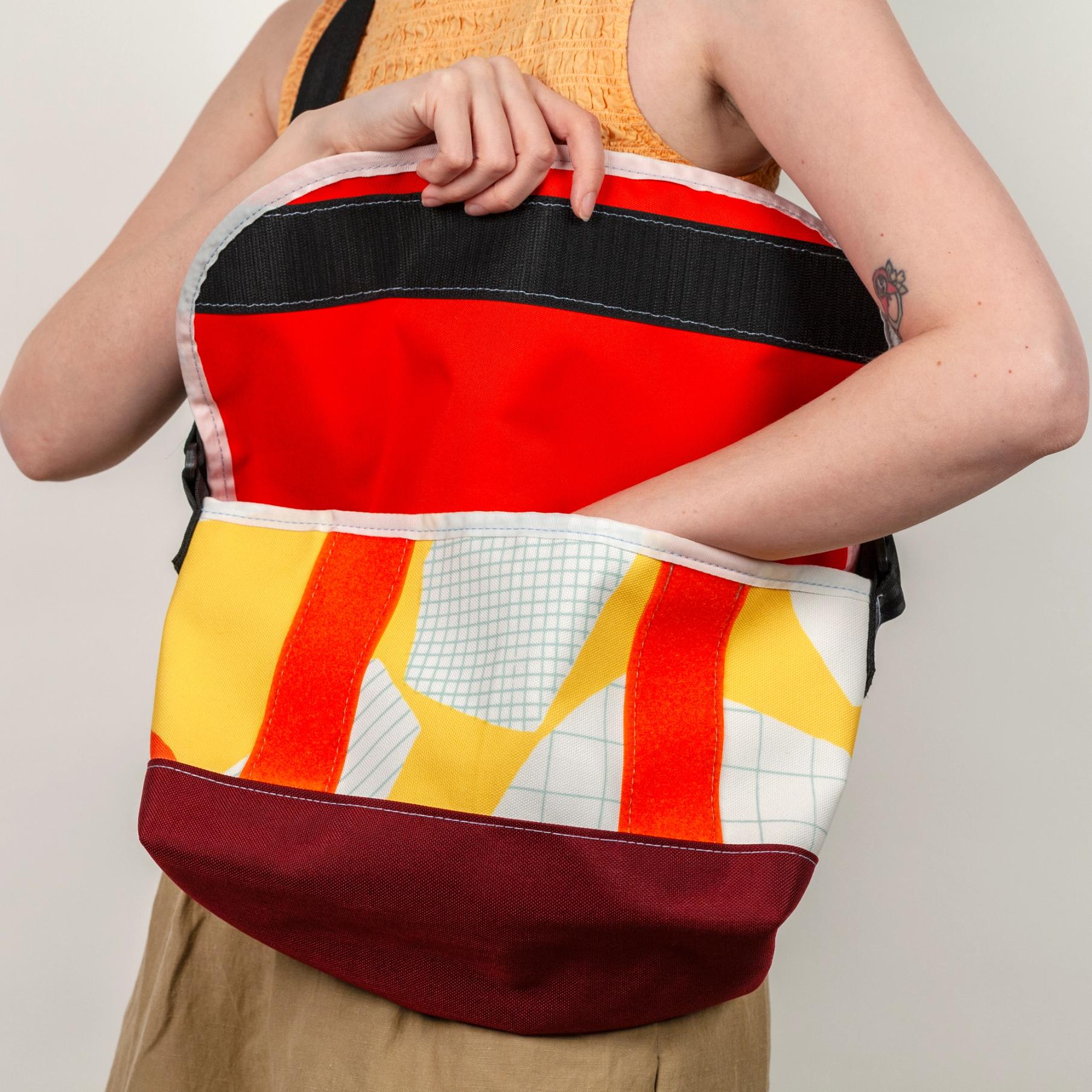 A model wearing a shallow, wide, vibrant yellow bag with bold abstract shapes (bright orange, white, white with light blue stripes, and white with a light blue grid pattern). The model is holding open the top flap which has a bright orange lining and black velcro closure with a white seam along the flap. The bag has a black strap and the base of the bag is solid maroon.