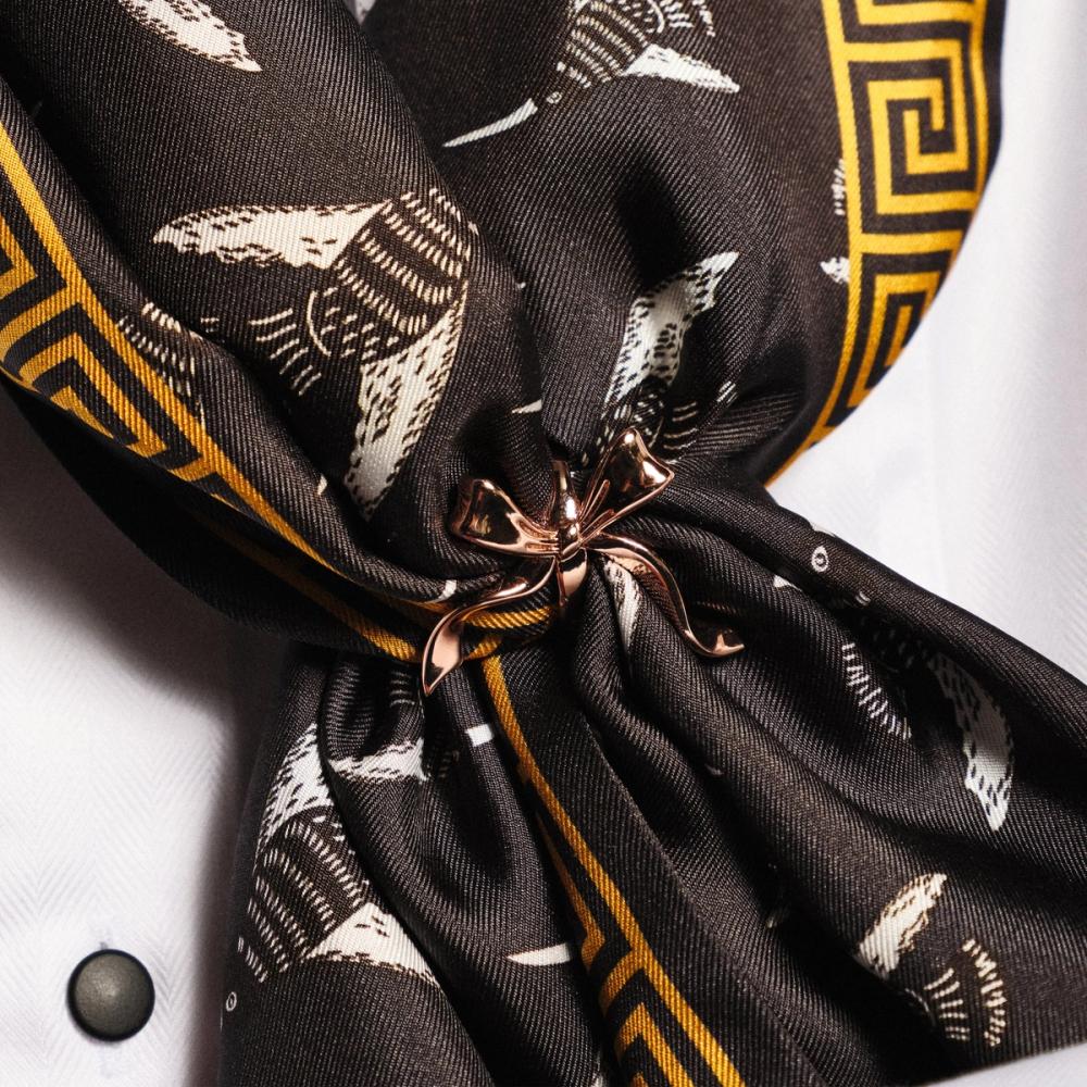 A rose gold colored scarf clip with a decorative bow pinches a black scarf together.