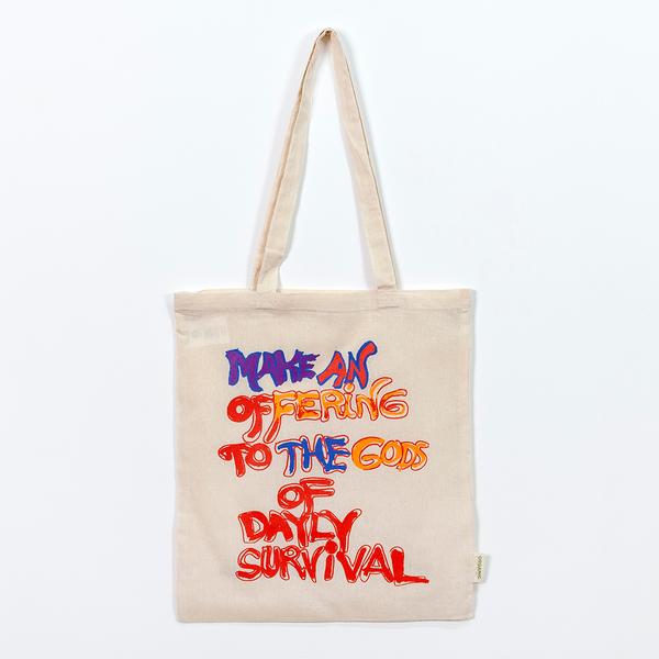 A photo of a cream-colored tote that reads in a hand-drawn message, "Make An Offering to the Gods of Daily Survival." The word daily is spelled d-a-y-l-y. Half the words appear in red while others are colored in with purple, pink, burnt yellow, and blue.