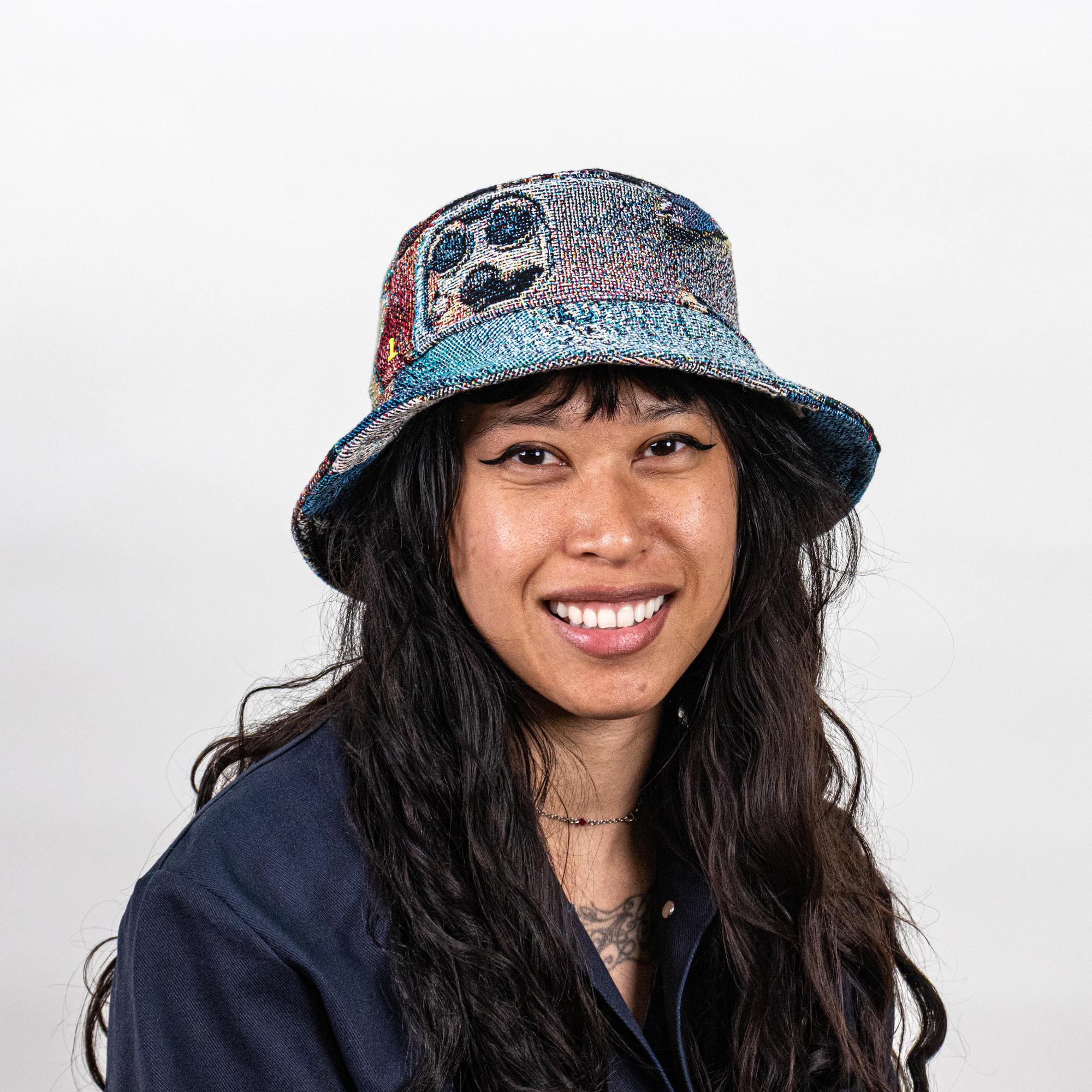 A model wearing a jacquard-woven bucket hat depicting textures, colors, and symbols of computers and tech. This view specifically features a back camera of an iPhone on the side of the bucket hat.