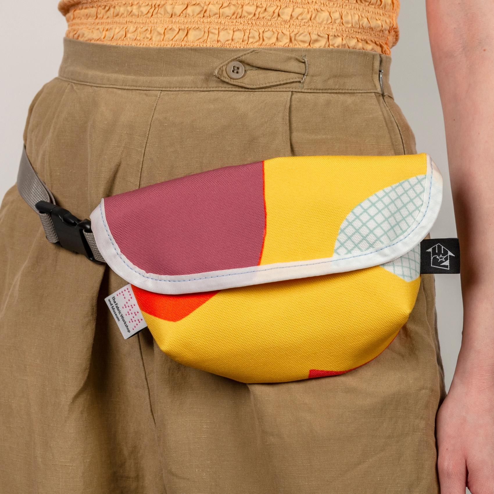 A model wearing a short, wide, vibrant yellow bag with bold abstract shapes (bright orange, purple, and white with a light blue grid pattern) on her hip (like a fanny pack). The bag has a velcro top flap closure with a white seam along the flap. The bag has a shiny gray strap with a black plastic buckle and two tags; one on the left and one on the right. The tag on the left is white with a pink dotted logo reading "FWM" with "The Fabric Workshop and Museum" written underneath. The tag on the right is black with an arrow. Inside the arrow are the letters "RE"