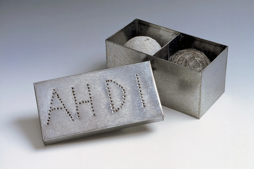 A photo of an opened tin box with the lid leaning against it against a gray background. The tin box is split into two cubes, each with a sphere inside. Inside the left cubicle the sphere appears light gray as if made of cement. Inside the right cubicle, the sphere is brown and appears to be made of a fibrous material. The lid, which faces the viewer, has the initials "AH" and "DI" inscribed with a series of small punctures made in the form of the letters.