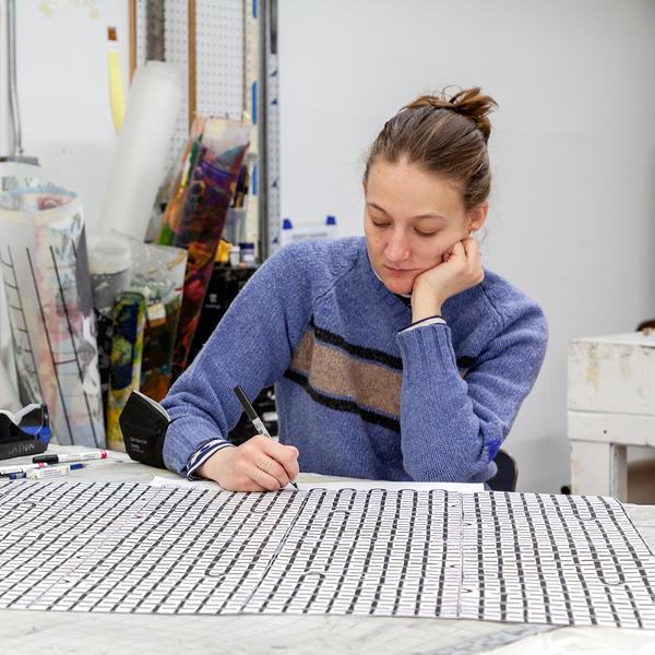 A photograph of a young white woman with light brown hair drawing while seated at a table. She is wearing a periwinkle colored sweater with tan and black stripes on the chest. She draws with a marker in her right hand while her left arm rests on the table with her hand supporting her head. The drawing features a repeated pattern of a loopy grid. Various rolls of materials stand upright in the background over her right shoulder.