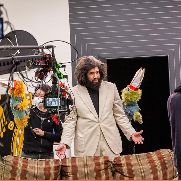 A behind-the-scenes photograph taken on a film set. A camera and rig obscures the left half of the image. At the center is a man wearing a cream suit with palms turned outward. To his left and right are two small puppets, including a puppeteer. He is standing in front of a grey wall with thin white stripes and a black door and behind a tan plaid couch.