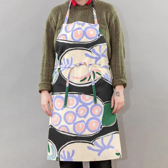 The same person is wearing the apron while standing against a grey background—this time, the apron is cinched around their waist and tied in a knot in the front.