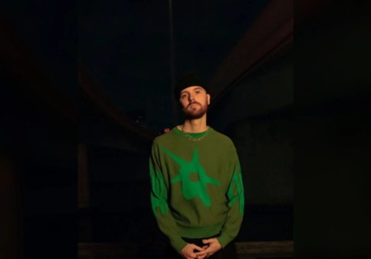 Tentendo standing in a dark studio portrait wearing a green patterned sweater