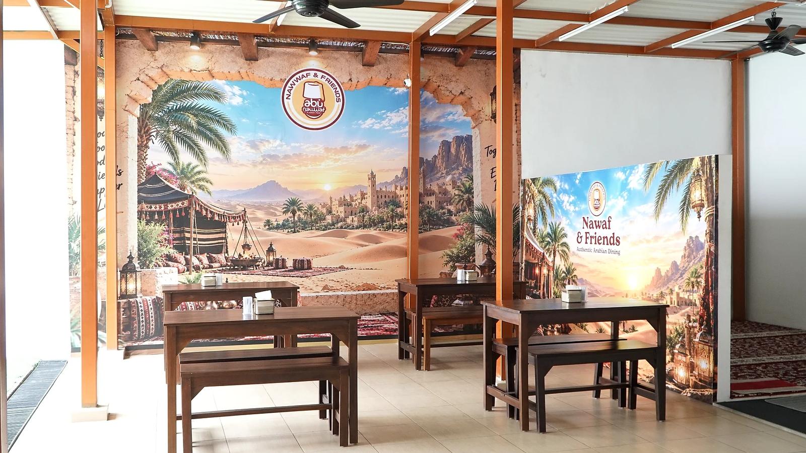 Area makan Nawwaf & Friends dengan meja dan bangku kayu, tiang oranye, dan dinding mural gurun bertuliskan logo Abu Nawwaf
