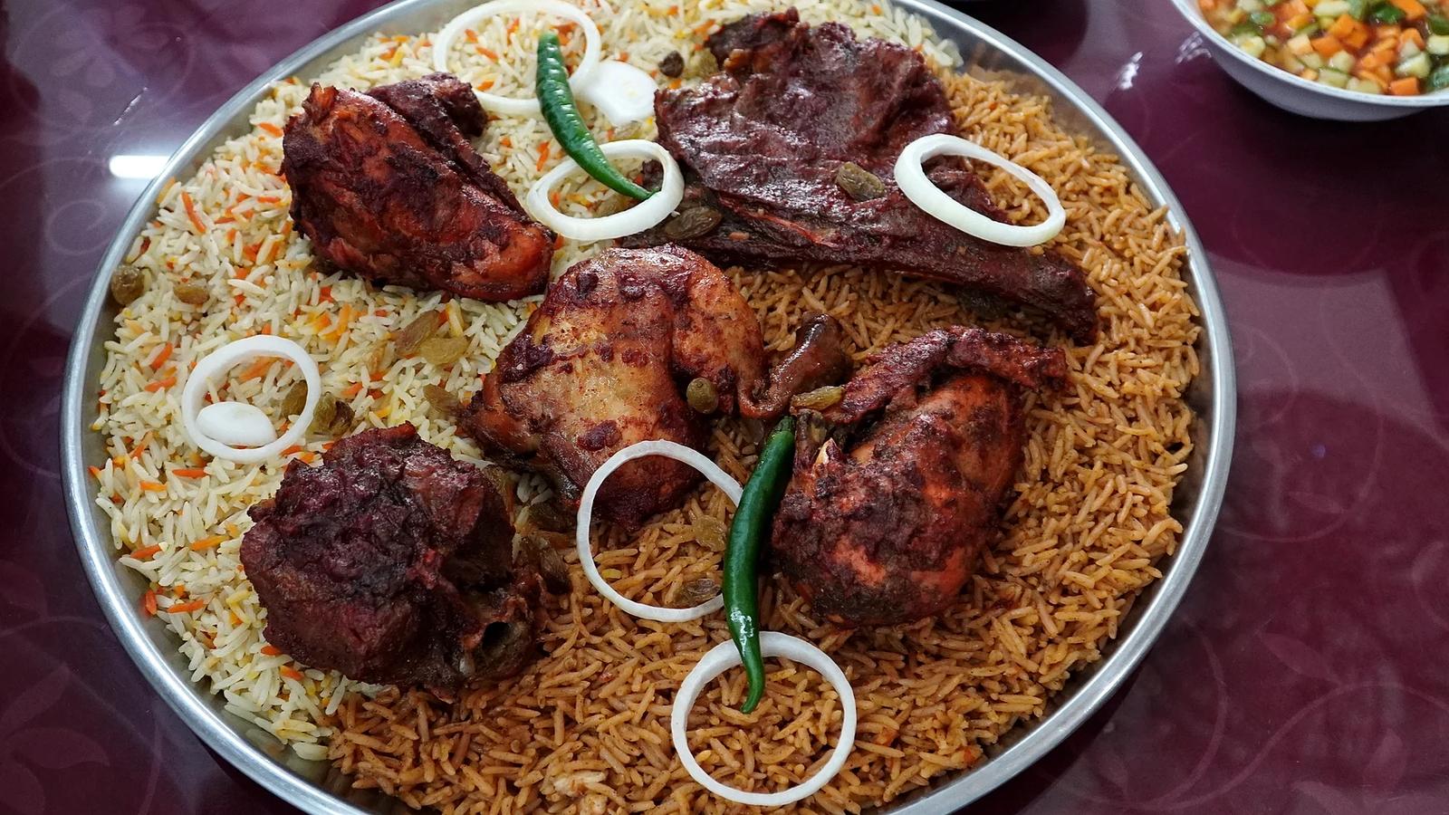 Nampan besar berisi nasi mandhi dan nasi kebuli dengan potongan ayam dan kambing, irisan bawang bombay, dan cabai hijau di atas meja ungu, dengan mangkuk sambal dan acar di sampingnya