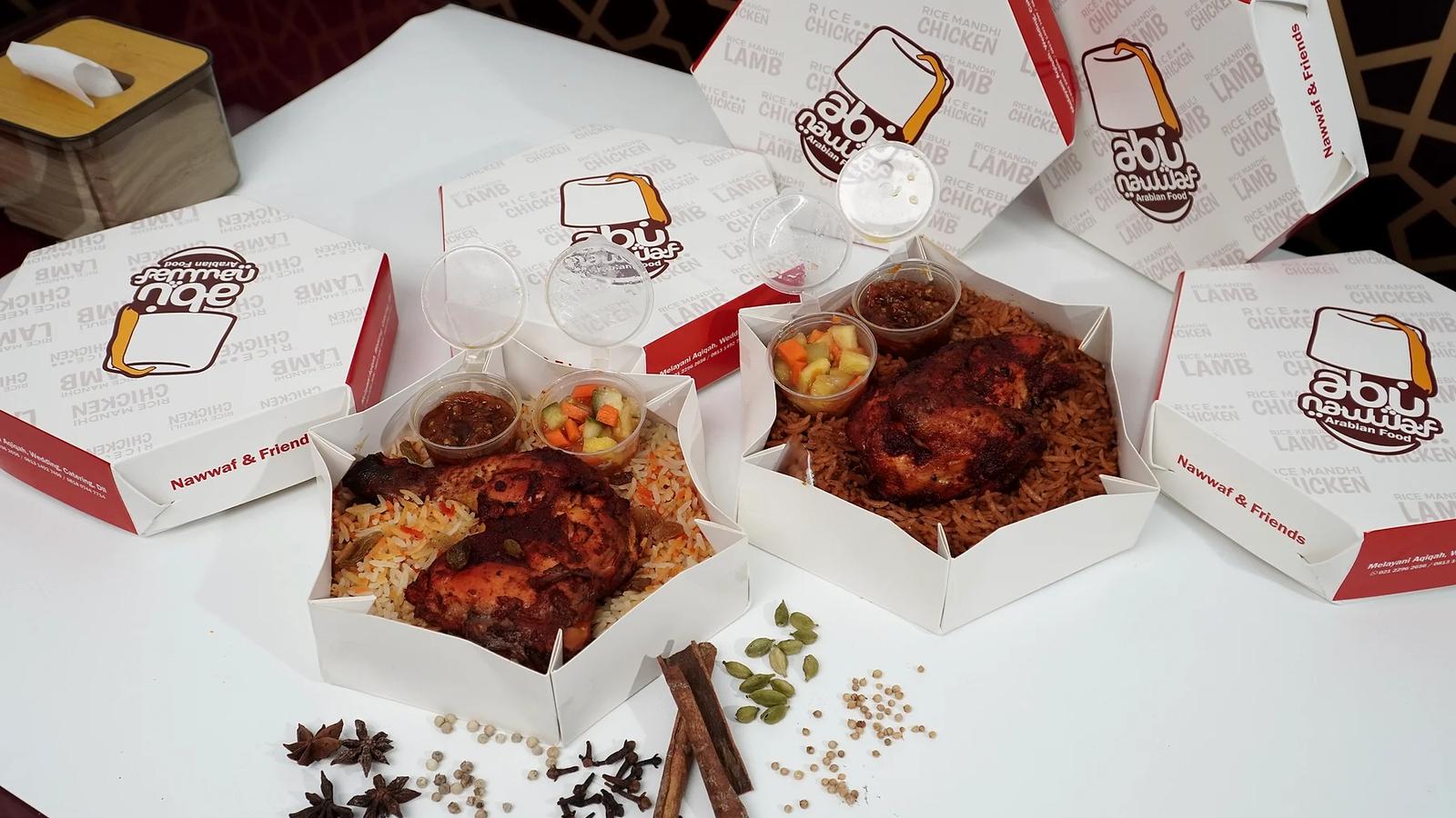 Box hexagon berisi nasi berbumbu dengan lauk ayam, sambal, dan acar, dikelilingi rempah di atas meja putih