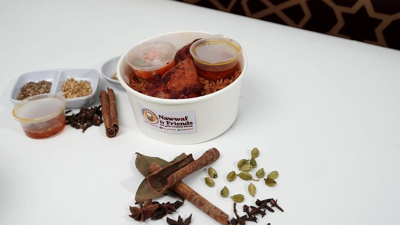 Rice bowl nasi berbumbu dengan satu potong ayam panggang, sambal dan acar, berlabel Nawwaf & Friends, dikelilingi rempah di meja putih