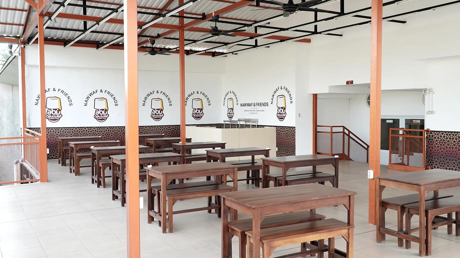Area makan lantai atas Nawwaf & Friends dengan deretan meja dan bangku kayu, tiang oranye, kipas angin plafon, dan dinding putih berlogo Abu Nawwaf