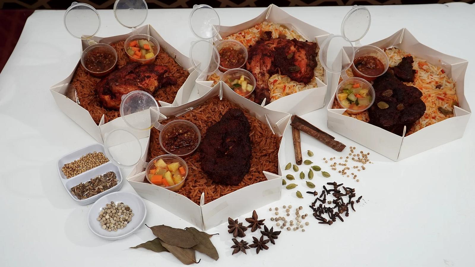 Empat box hexagon terbuka berisi nasi mandhi dan kebuli dengan ayam dan kambing, sambal, dan acar, dikelilingi rempah di meja putih