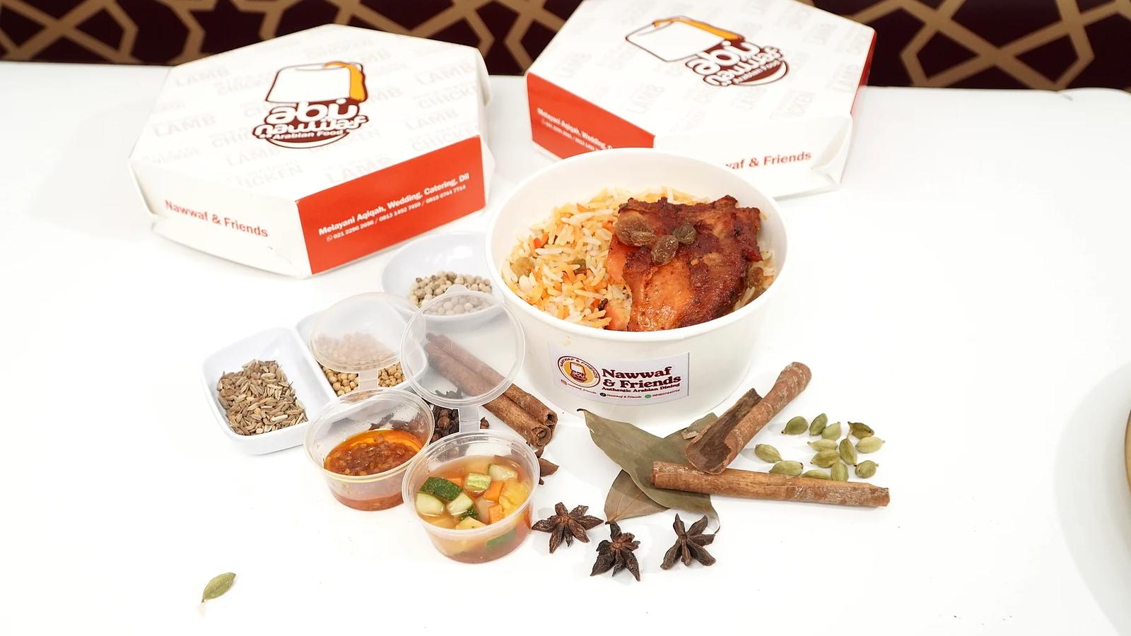 Rice bowl nasi berbumbu dengan lauk panggang, sambal, dan acar di meja putih bersama rempah, dengan dua box Abu Nawwaf bertuliskan Melayani Aqiqah di belakangnya