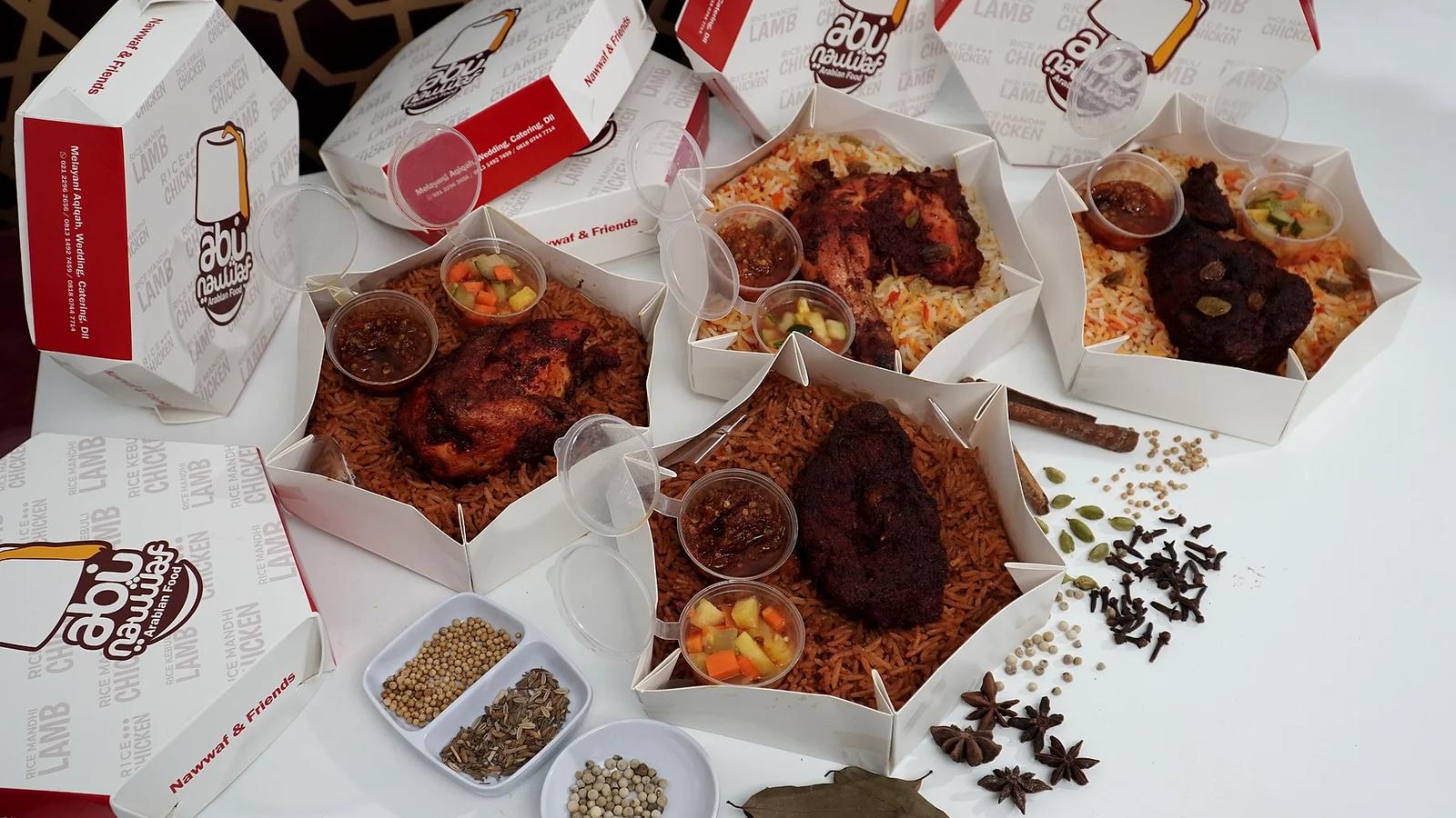 Beberapa kotak hexagon Abu Nawwaf terbuka berisi nasi berbumbu dengan ayam dan daging panggang, sambal dan acar dalam cup, dikelilingi rempah seperti kapulaga, cengkih, dan bunga lawang di meja putih