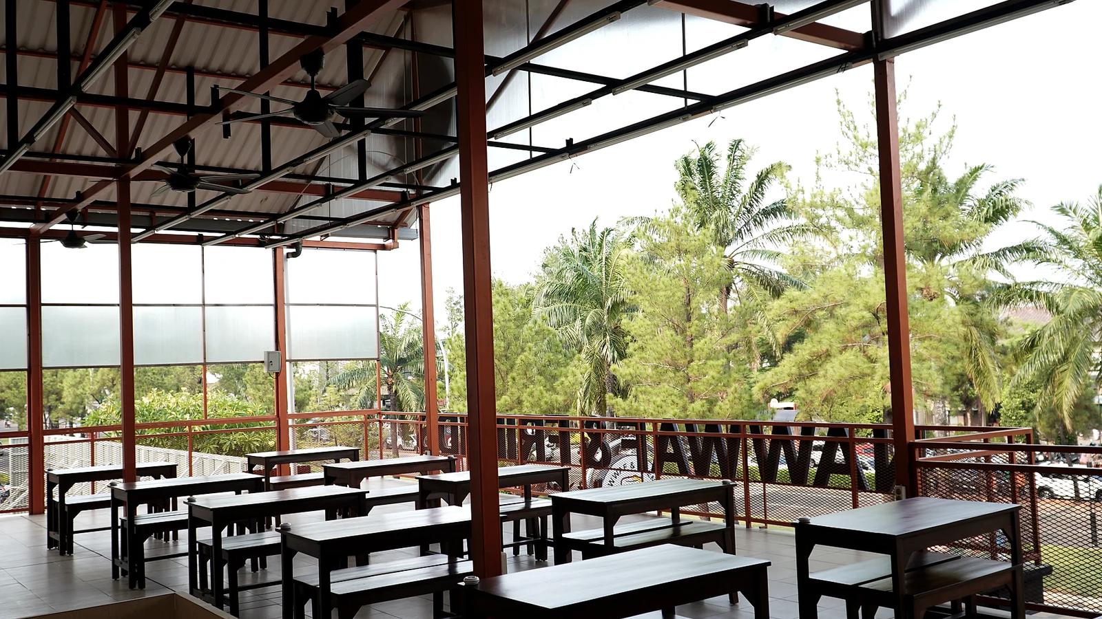 Area makan semi-outdoor di lantai atas Nawwaf & Friends dengan deretan meja dan bangku kayu, kipas angin di langit-langit, pagar bertuliskan Nawwaf & Friends, dan pepohonan palem di luar