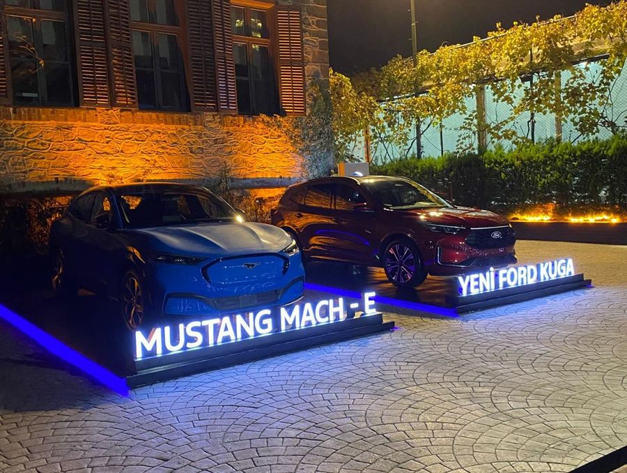 Mustang Mach-E and new Ford Kuga on raised platforms with illuminated model lettering, in a cobbled courtyard with orange uplighting on the stone façade after dark - BACKSTAGE Event production