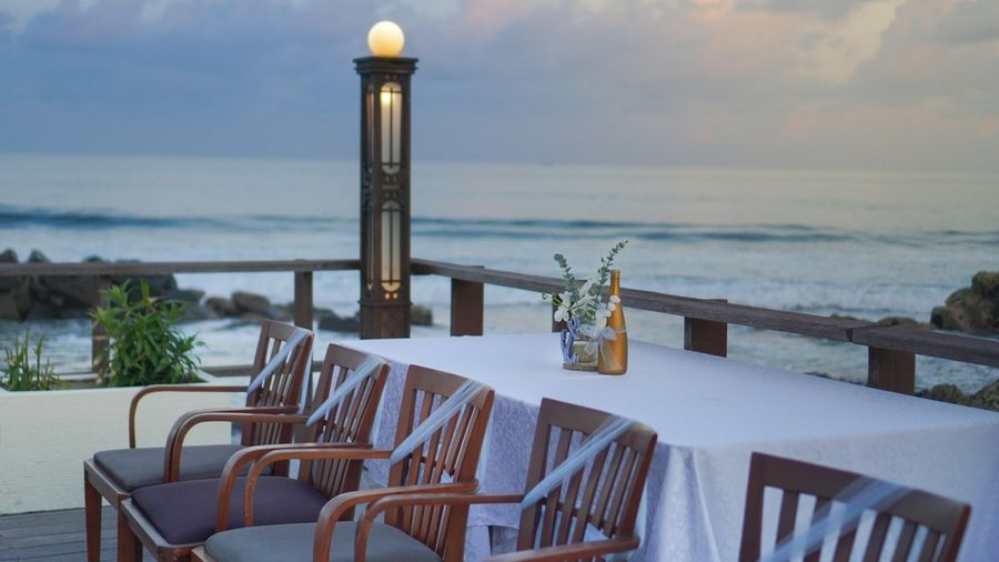 Lange Tafel auf einem Holzdeck am Wasser in der Dämmerung, Laternenlicht und dahinter das Meer