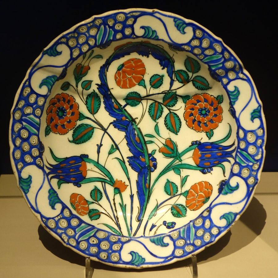 White ceramic dish with a blue border, painted with blue tulips, orange-red round flowers and green leaves, on a display stand