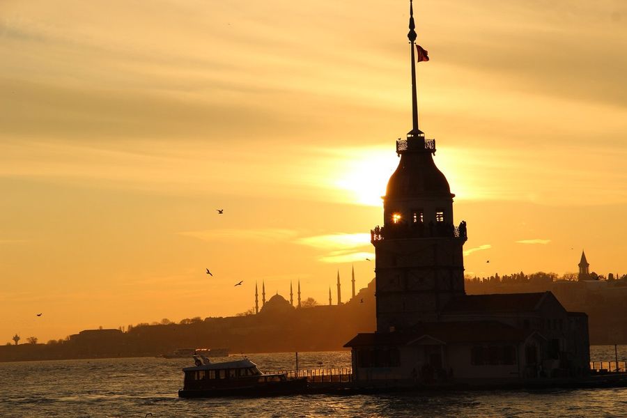 Maiden's Tower silhouetted against an orange sunset on the Bosphorus, with minarets on the far shore and a small boat passing