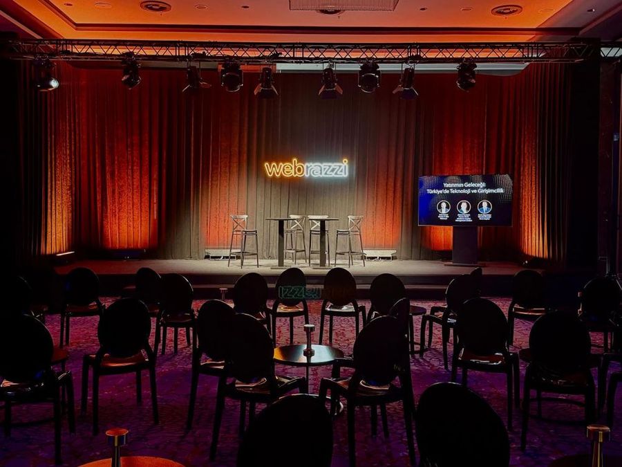 Conference stage with orange-lit curtains under a truss lighting grid, a neon Webrazzi logo, a session screen and high tables, with rows of chairs facing it, at the Webrazzi summit - BACKSTAGE Event production