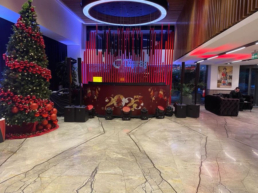 Lobby stage for Tüpraş's new-year celebration, with a red slatted wall carrying the company logo, speakers, moving-head lights and a decorated tree - BACKSTAGE Event production