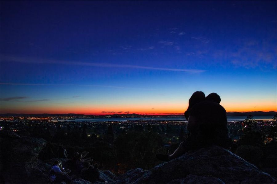 A couple standing on a hillside at dusk, looking out over distant city lights
